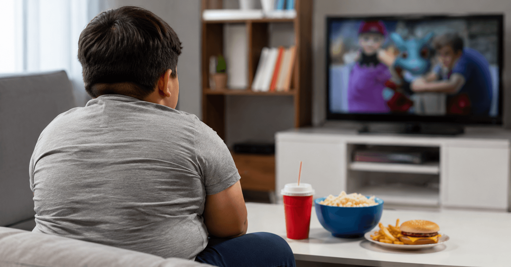 Imagem ilustrativa de uma criança com excesso de peso vista de costas enquanto assiste televisão, com alimentos sobre a mesa à sua frente. A fotografia representa um cenário comum associado ao sedentarismo e à obesidade infantil, tema que reforça a importância de hábitos saudáveis, alimentação equilibrada e prática regular de atividades físicas desde a infância.